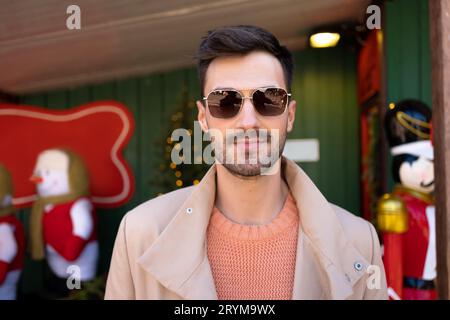 Uomo vicino alle attrazioni natalizie all'aperto. Si trova accanto a una casa di legno decorata per Natale. Bel ragazzo con occhiali da sole e trench che posa con decorazioni di Capodanno sullo sfondo Foto Stock