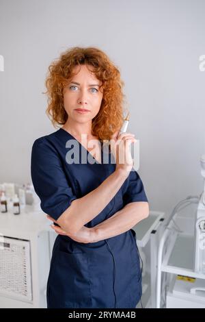 Cosmetologa femminile presso lo studio medico. Dermatologo donna rossa che indossa scrub tiene una fiala all'ospedale. Medico di cura della pelle in uniforme medica blu navy sta in studio di clinica estetica Foto Stock