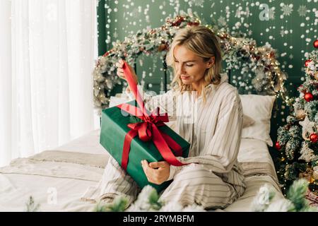 Una felice donna sorridente in pigiama seduta sul letto sgancia il nastro con un regalo di Natale. Concetto festivo di holida di Capodanno Foto Stock