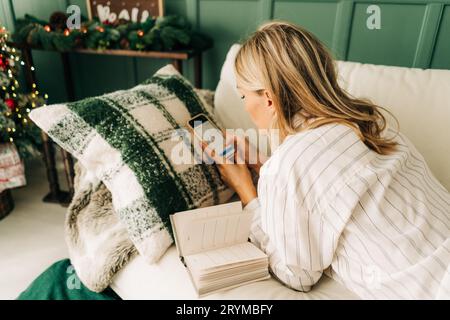 Vista posteriore di una donna d'affari bionda e impegnata sdraiata sul divano a casa, concentrata sullo smartphone e alla ricerca di scambi online. Investire fina Foto Stock