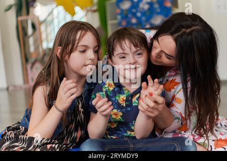 Una ragazza e una donna abbracciano un bambino con sindrome di Down in una moderna istituzione prescolare Foto Stock