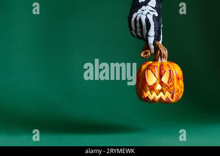 Le mani con guanti scheletro mantengono la zucca in ceramica jack-o'-lantern su sfondo verde Foto Stock