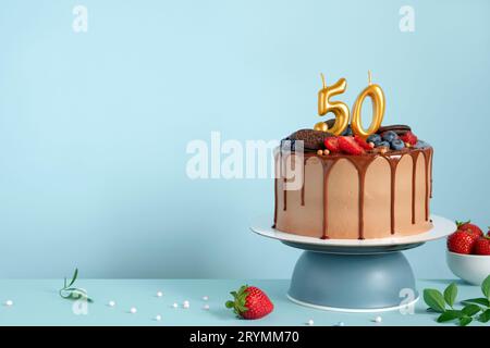 Torta di compleanno al cioccolato con bacche, biscotti e numero cinquanta candele dorate su sfondo blu parete, spazio copia Foto Stock