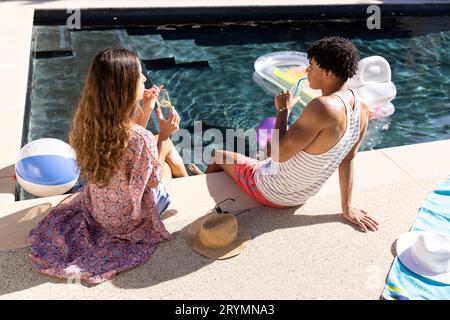 Vista posteriore di coppia varia Happy FIT bere cocktail seduti a bordo piscina al sole Foto Stock