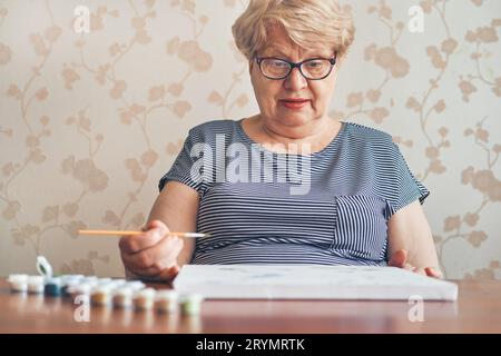 Attraente vecchia donna che dipinge per numeri a casa. Hobby e arte terapeutica ispirati. Messa a fuoco selettiva Foto Stock