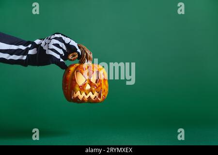 Le mani con guanti scheletro mantengono la zucca in ceramica jack-o'-lantern su sfondo verde Foto Stock