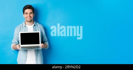 Bello giovane uomo introdurre il prodotto sullo schermo del laptop, mostrando il computer e sorridendo, in piedi su sfondo blu Foto Stock