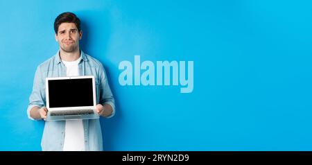 Un ragazzo oscuro e dispiaciuto che mostra lo schermo del notebook e la grimacing, avendo dubbi, in piedi su sfondo blu in casual Foto Stock