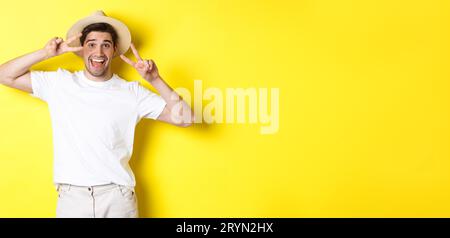 Concetto di turismo e vacanza. Un turista felice in posa per una foto con i segni della pace, sorridente eccitato, in piedi contro la b gialla Foto Stock