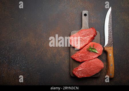 Tre bistecche di manzo crude su tagliere di legno con rosmarino su tagliere di legno con coltello su buio Foto Stock