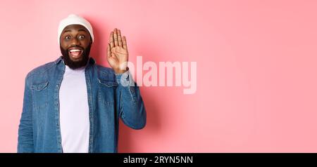 Amico sorridente uomo nero che ciao, con la mano, vi accoglie, in piedi su sfondo rosa Foto Stock