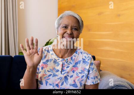 Donna biracial anziana felice che fa la videochiamata sorridendo e ondeggiando seduto sul divano a casa Foto Stock