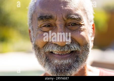 Ritratto di felice uomo biraciale anziano con barba che guarda la macchina fotografica e sorridente in giardino soleggiato Foto Stock