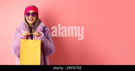 Donna anziana asiatica alla moda che va a fare shopping con occhiali da sole e abiti invernali alla moda, tenendo in mano un sacchetto di carta dal negozio e sorridendo Foto Stock
