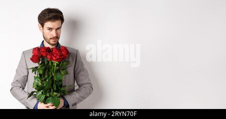 Bellissimo macho che va in giro con la tuta, con bouquet di rose rosse e sorridendo davanti alla macchina fotografica, facendo gif a sorpresa per il giorno dei valentini Foto Stock