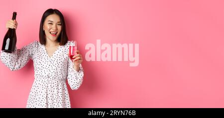 Felice donna asiatica che festeggia, divertendosi e festeggiando, versando un bicchiere di champagne e ridendo, in piedi su sfondo rosa Foto Stock