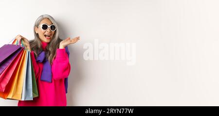 Donna asiatica anziana ed elegante in occhiali da sole che va a fare shopping in vendita per le vacanze, tenendo borse di carta e sorridendo, in piedi su bianco bac Foto Stock
