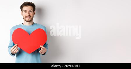 Concetto di relazione e amore. Bell'uomo caucasico in maglione che regge grandi valentine rosse giorno cuore ritaglio e sorridente, confes Foto Stock