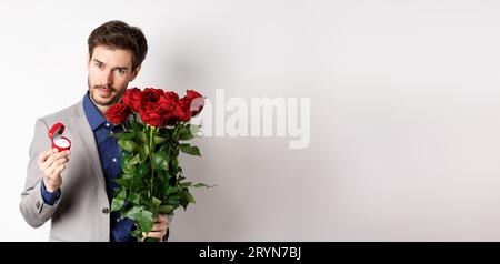 Ragazzo romantico che mostra bouquet di rose rosse, puntando ai fiori e in  piedi vicino ai palloncini cuori il giorno di San Valentino, preparare regali  per il suo amante, bianco Foto stock 