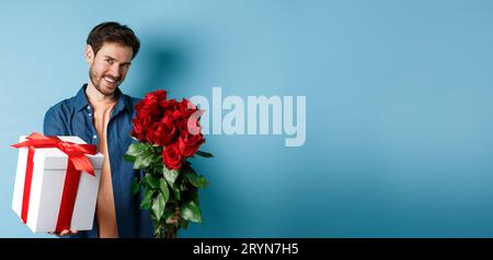 Concetto Love and Valentines Day. Romantico uomo sorridente che vi regala una scatola regalo e un bouquet di fiori in data, in piedi sopra il bac blu Foto Stock