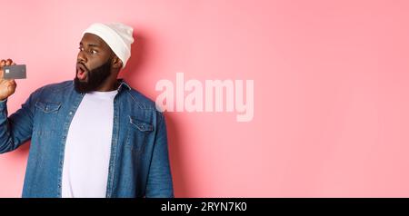 Concetto di shopping. Immagine di un uomo afro-americano bearded che staring scioccato alla carta di credito, in piedi contro sfondo rosa Foto Stock
