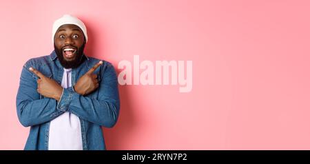 Felice ragazzo afro-americano hipster che punta le dita lateralmente, sorride e mostra due scelte, mostra offerte, in piedi ov Foto Stock