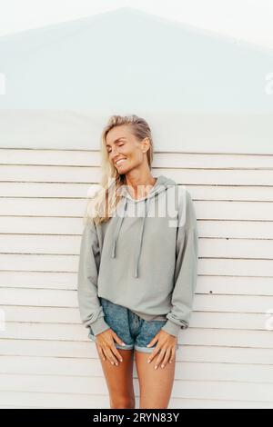 Bella bella donna bionda abbronzata con pantaloncini in denim e un'elegante felpa con cappuccio che si diverte e ride sinceramente. Foto Stock