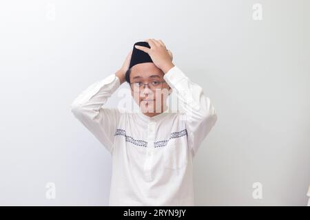 Ritratto di un giovane musulmano asiatico che cerca di regolare il suo songkok o Skullcap nero. Immagine isolata su sfondo bianco Foto Stock