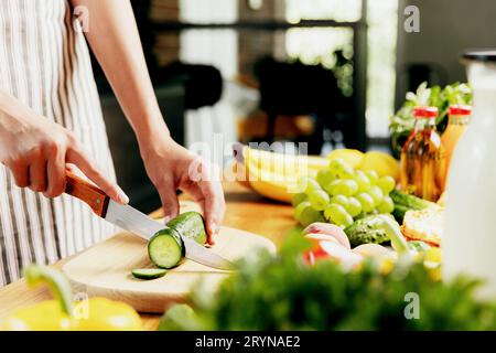 Primo piano sulle mani di una giovane casalinga o di una ragazza che affetta il cetriolo con un coltello da cucina, taglia il cibo sul tagliere di legno. Stacnello femminile irriconoscibile Foto Stock