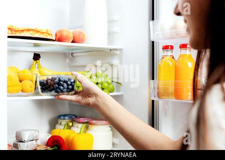 Donna che prende, afferra o preleva una scatola di mirtilli dal ripiano aperto del frigorifero o dal cassetto del frigorifero pieno di frutta, verdura, banana, pesche, Foto Stock