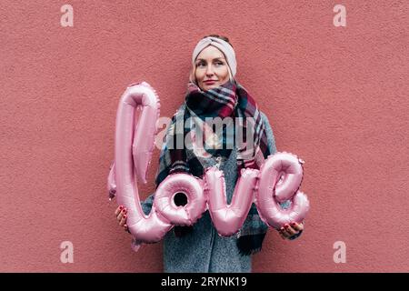 Piacevole giovane donna in un cappotto vicino alla parete rosa Tiene la parola gonfiata AMORE Foto Stock