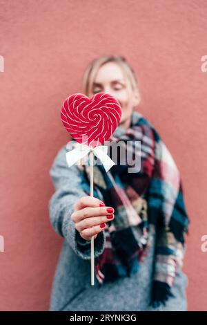 Primo piano di una donna che regge un lollipop di caramello a forma di cuore. Contenuto e data del giorno di San Valentino. Atmosfera romantica. Sia il mio Valentine. Foto Stock