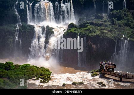 Vista ravvicinata delle spettacolari cascate con una lussureggiante vegetazione verde e una piattaforma per visitatori, Iguazu fa Foto Stock