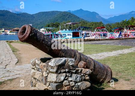 Primo piano di un vecchio cannone arrugginito di fronte a uno spazio aperto con erba, barche colorate e foresta pluviale Foto Stock