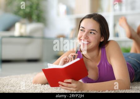 Donna felice che scrive all'ordine del giorno pensando di guardare un lato sdraiato su un tappeto a casa Foto Stock