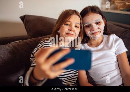 Due belle ragazze adolescenti con tessuto usa e getta idratante maschere di bellezza sui loro volti prendono un selfie. Carine amiche femminili lo fanno Foto Stock