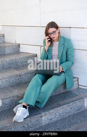 Donna d'affari seduta sulle scale fuori dall'edificio dell'ufficio mentre lavora con il computer portatile e sta telefonando Foto Stock