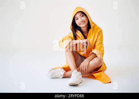 Sorridente ragazza asiatica con bretelle vestite con un impermeabile giallo brillante in posa con cappuccio sulla testa su sfondo bianco dello studio seduta sul pavimento con una copia di spumante Foto Stock