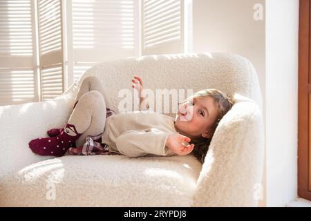 Simpatica bambina giace su una morbida sedia bianca, ingannando, facendo facce vicino all'albero di Natale. Bambino in un'atmosfera accogliente a casa. Interi festivi Foto Stock