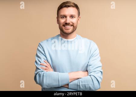 Ritratto di un uomo sorridente con le mani incrociate sul petto su sfondo beige che guarda la fotocamera. Foto Stock