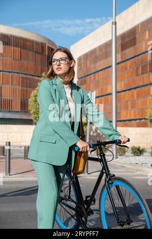 Elegante donna dai capelli rossi in costume verde che cammina in bicicletta nel quartiere finanziario con un edificio moderno sullo sfondo Foto Stock