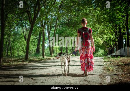 Cammina con un cucciolo una giovane donna in una gonna colorata e una t-shirt Bordeaux è accompagnata da una brillante giornata estiva di cane dalmata al guinzaglio in pennsylvania Foto Stock