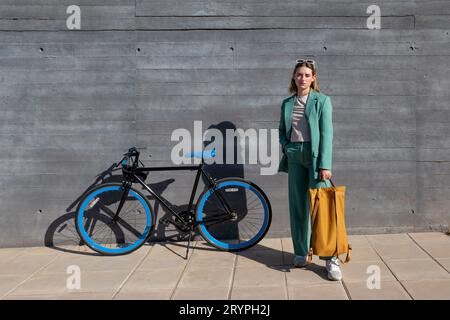 Elegante donna da lavoro a pelo di zenzero con tuta verde e zaino giallo in piedi con bicicletta su sfondo nero Foto Stock