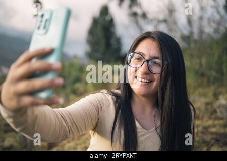 Donna ispanica con gli occhiali che scatta un selfie nella natura al tramonto Foto Stock