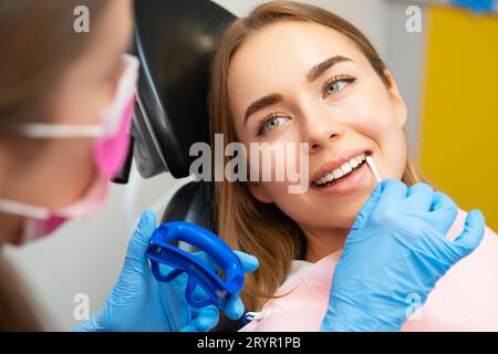 Il dentista applica il rilevatore di placche sui denti anteriori della donna paziente in occhiali protettivi. Il medico esegue la procedura per avere anche un sorriso Foto Stock