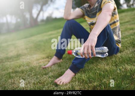 Giovane uomo caucasico corridore rilassante tenendo bottiglia di acqua potabile e. seduto sull'erba nel parco all'aperto dopo lo sport a. presto Foto Stock