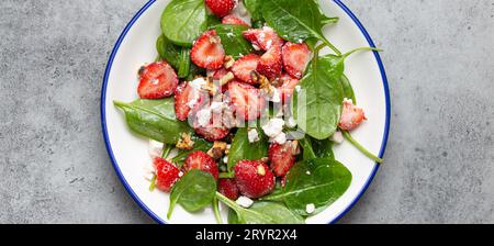 Insalata estiva leggera e salutare con fragole fresche, spinaci, crema di formaggio e noci su piatto di ceramica bianca, sfondo in pietra rustica grigia Vista dall'alto. Foto Stock