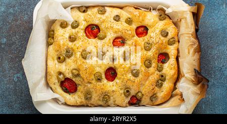 Testa di focaccia tradizionale italiana con olive verdi, olio d'oliva, pomodori ciliegini e rosmarino in teglia da forno sul rustico scuro Foto Stock