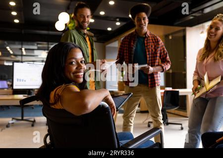 Felice donna afro-americana in sedia a rotelle con diversi colleghi durante una riunione informale in ufficio Foto Stock