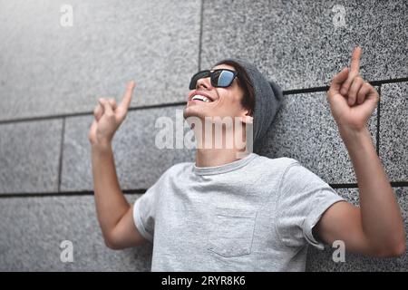Bel giovane con gli occhiali da sole sta puntando, guardando in alto e sorridendo, in piedi contro il muro grigio. Foto Stock